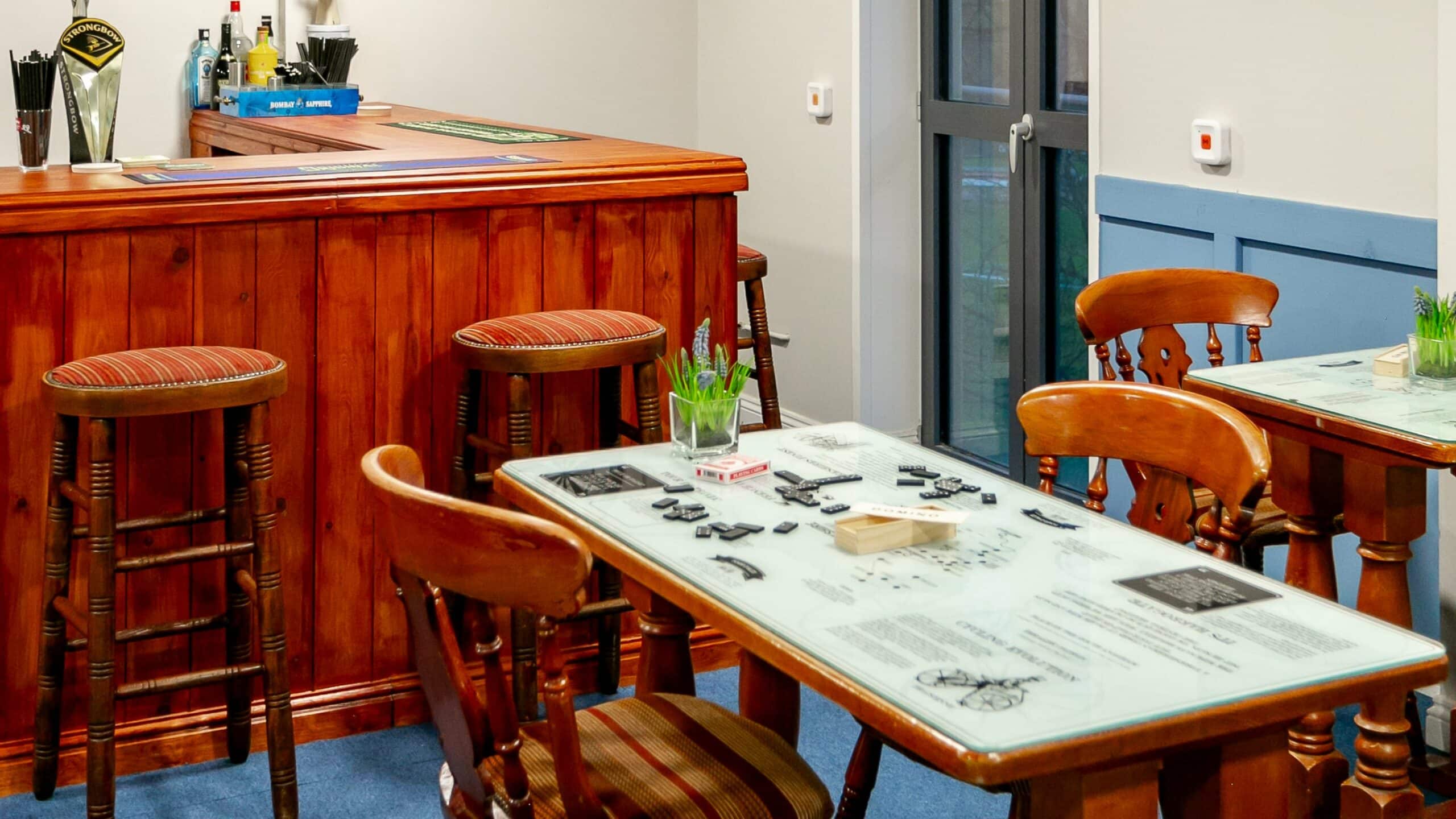 Pub-style bar with a wooden counter, bar stools and a table set with dominoes at Bridge House Care Home, Brighouse