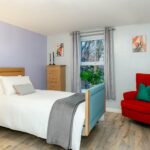 Lilac bedroom with oak furniture, a red armchair and a window with grey curtains at Bridge House Care Home, Brighouse