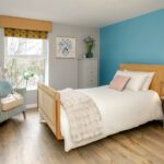 Bedroom with a teal feature wall, wooden-framed bed, armchair and tree-view window at Bridge House Care Home, Brighouse