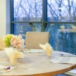 Round dining table set with napkins and flowers beside floor-to-ceiling windows at Bridge House Care Home, Brighouse