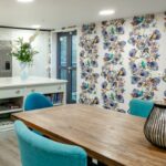 Kitchenette with teal dining chairs, a long wooden table and bird-print wallpaper at Bridge House Care Home, Brighouse