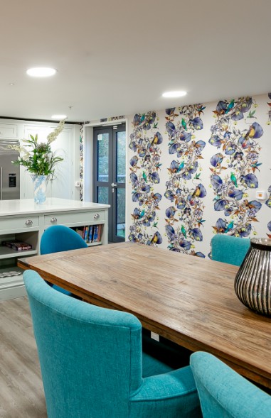 Kitchenette with teal dining chairs, a long wooden table and bird-print wallpaper at Bridge House Care Home, Brighouse