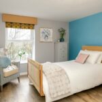 Bedroom with a teal feature wall, made-up bed, armchair and window overlooking trees at Bridge House Care Home, Brighouse