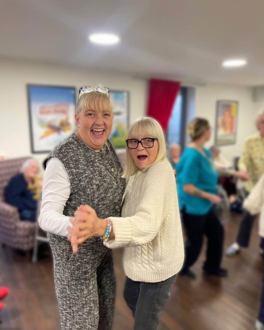 Two women laughing and dancing hand in hand in a lounge at Whitby Court Care Home, Whitby