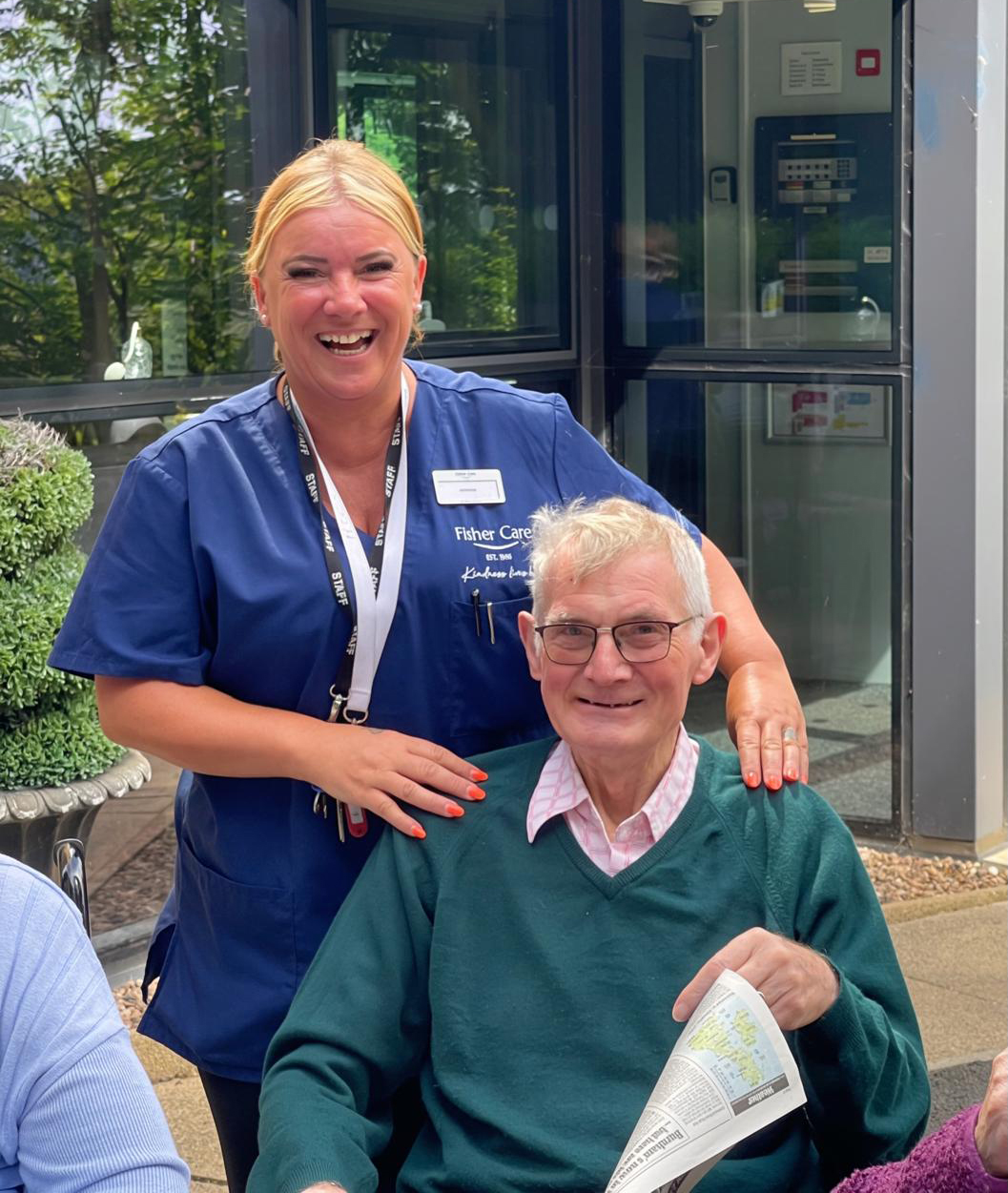 Smiling carer in Fisher Care uniform with her hands on the shoulders of a seated resident holding a newspaper