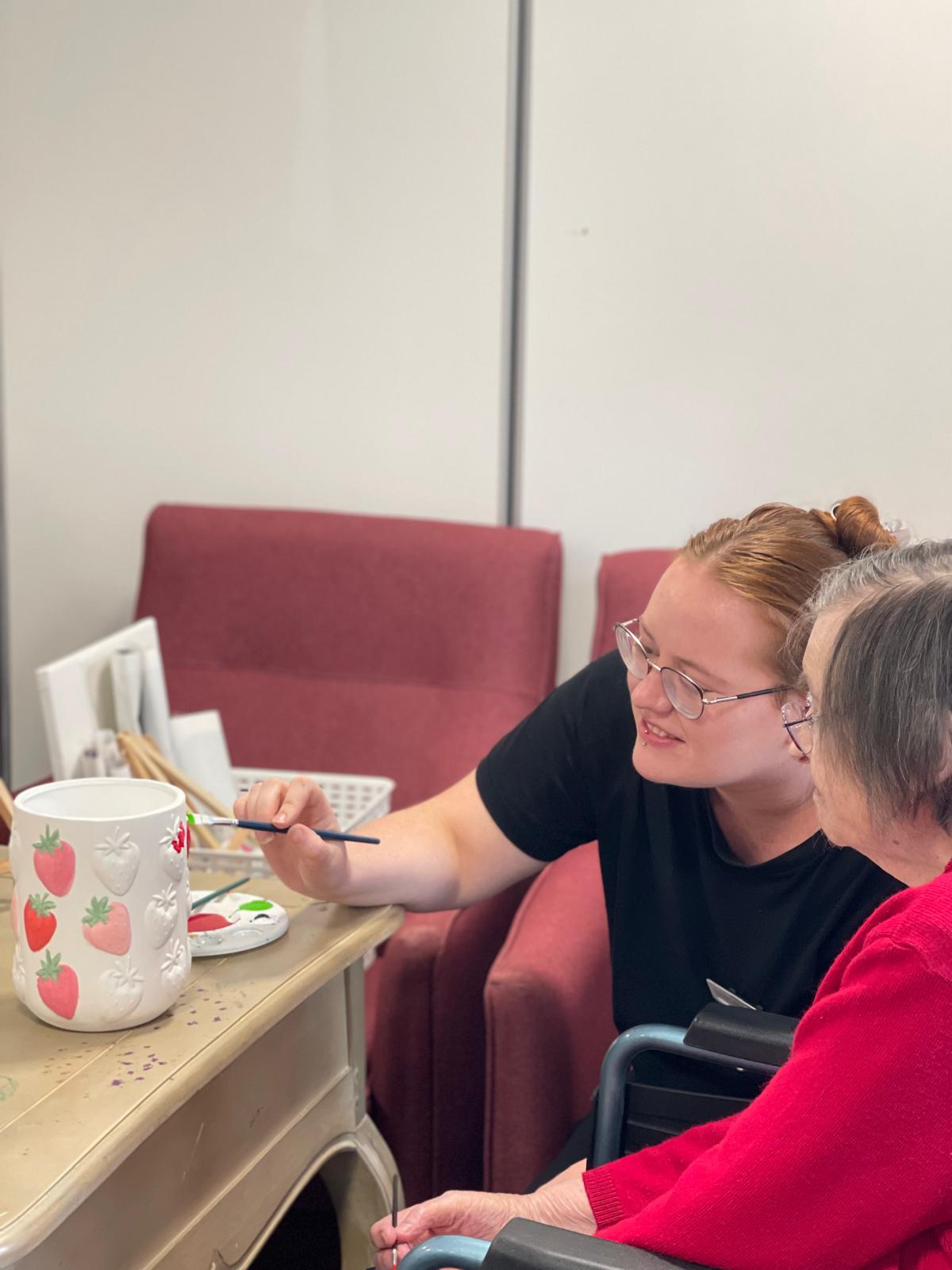 A resident and a young woman painting a strawberry-patterned ceramic pot together