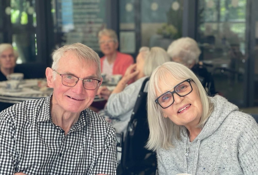 Smiling older man and woman sitting side by side at a table, with others enjoying tea in the background