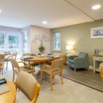 Bright dining room with laid round tables, garden windows and an armchair by a lamp at Whitby Court Care Home, Whitby