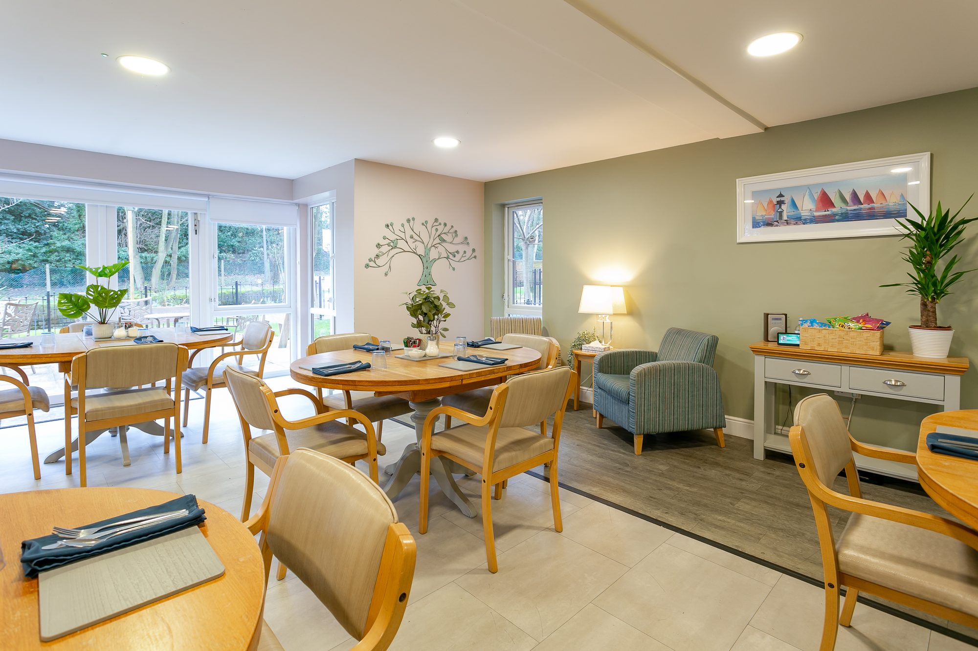 Bright dining room with laid round tables, garden windows and an armchair by a lamp at Whitby Court Care Home, Whitby