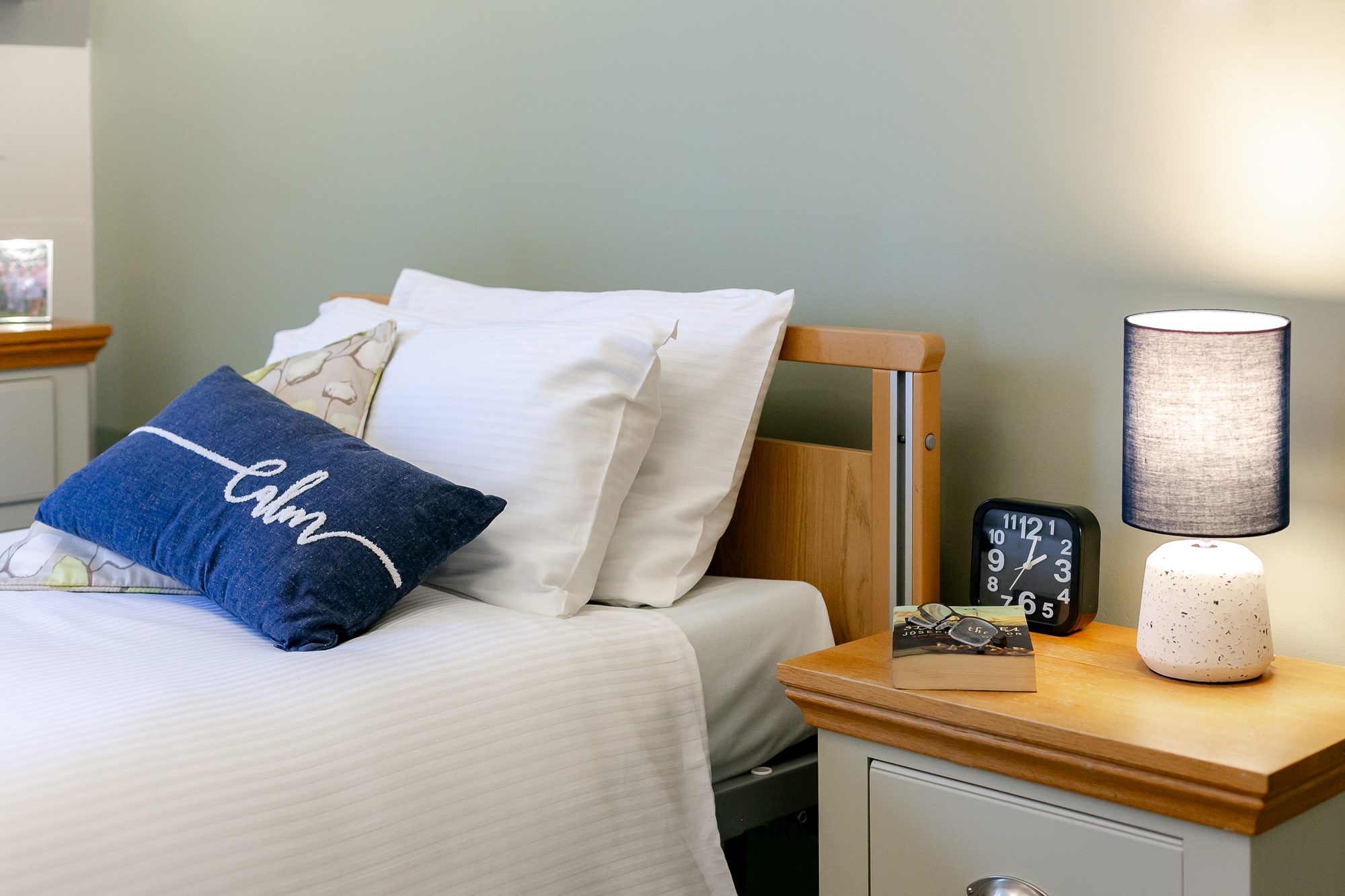 Bedside table with a book, reading glasses, clock and lit lamp at Whitby Court Care Home, Whitby