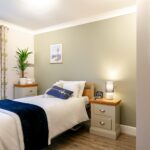 Bedroom with a navy throw on the bed, lit bedside lamp and framed coastal prints at Whitby Court Care Home, Whitby