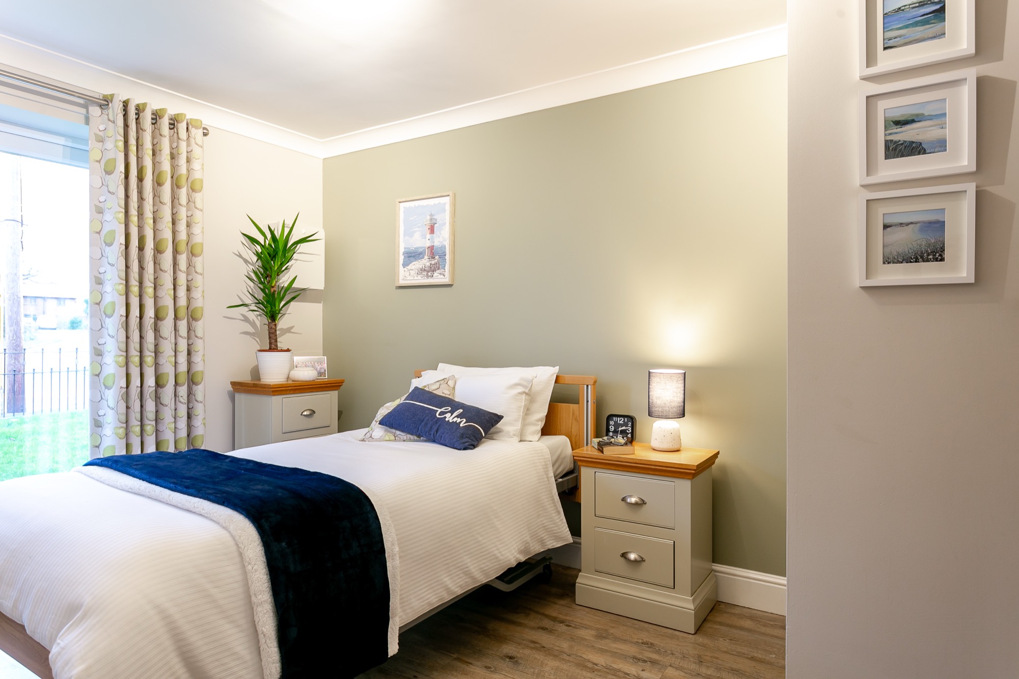 Bedroom with a navy throw on the bed, lit bedside lamp and framed coastal prints at Whitby Court Care Home, Whitby
