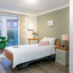 Bedroom with adjustable bed, pink bedside lamp and patterned curtains framing patio doors at Whitby Court Care Home, Whitby