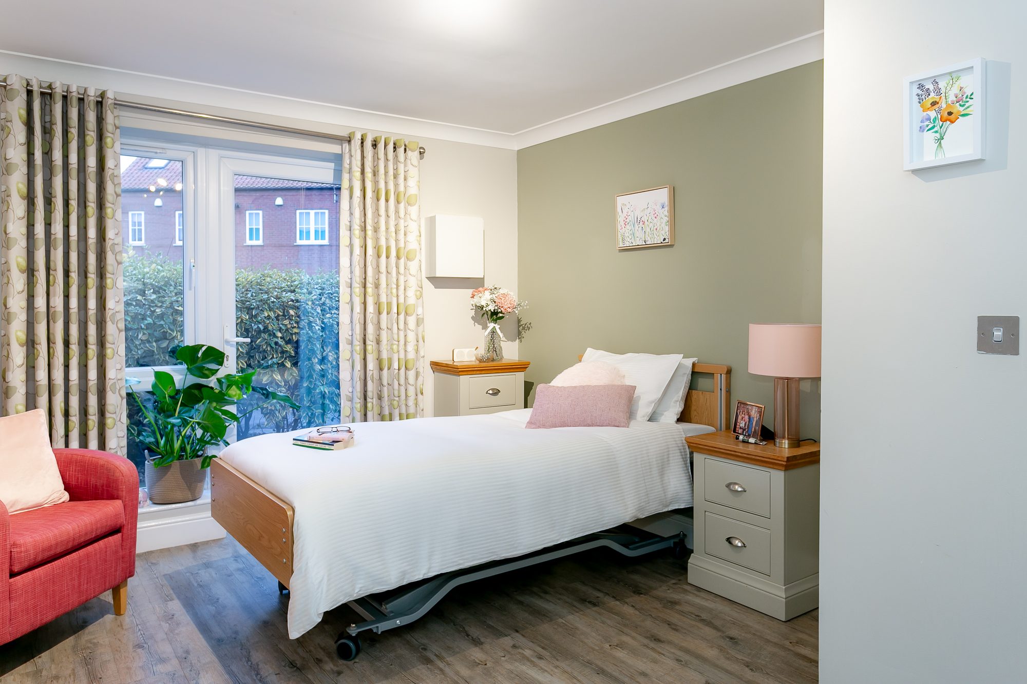 Bedroom with adjustable bed, pink bedside lamp and patterned curtains framing patio doors at Whitby Court Care Home, Whitby